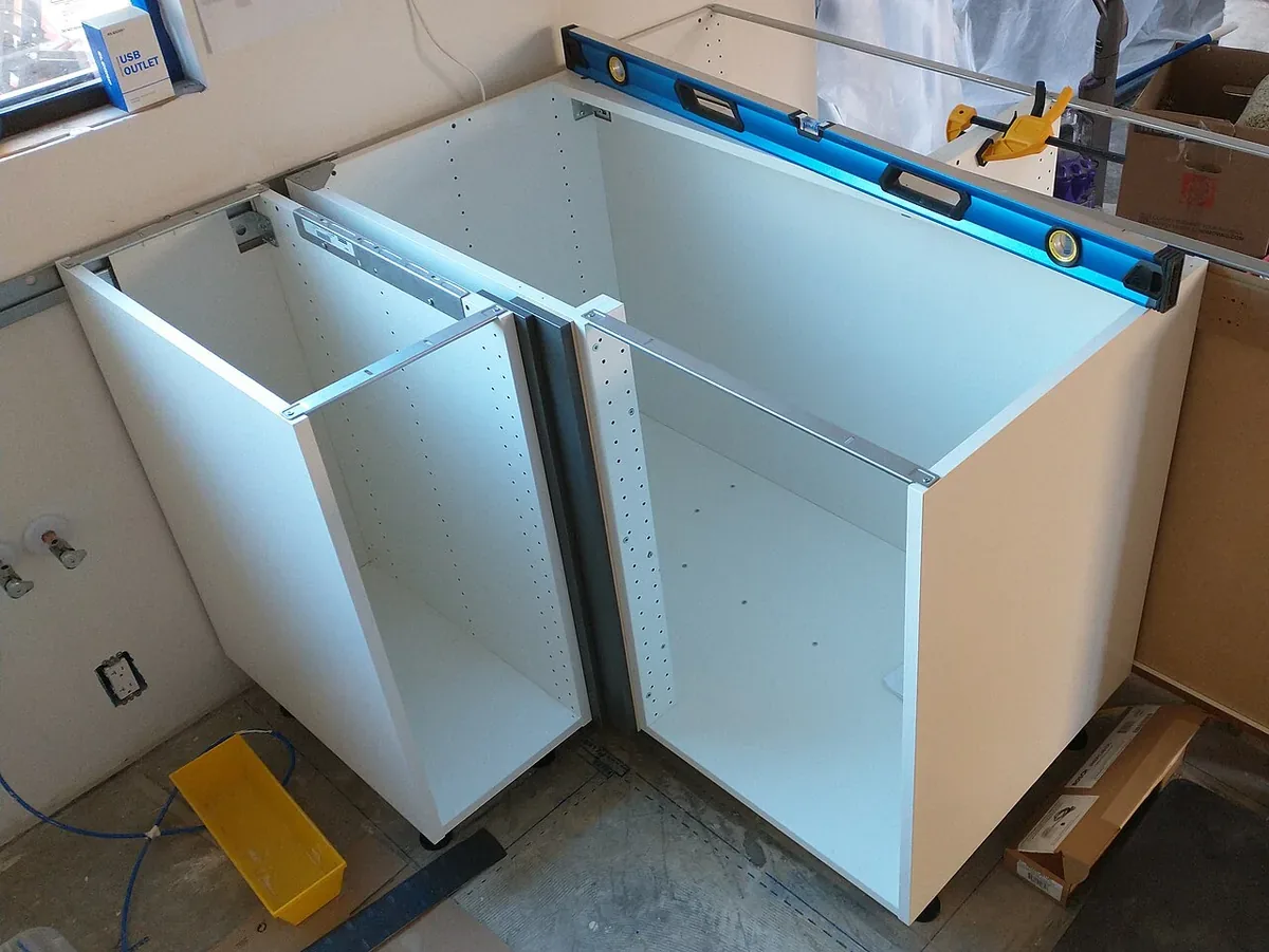 Two base cabinet boxes being set into a corner during a kitchen cabinet installation, with a spirit level across the top and drawer slides fitted
