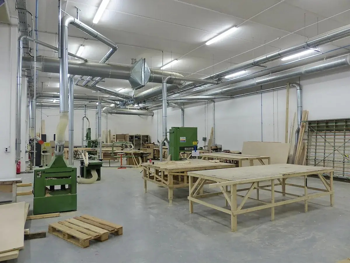 Cabinet shop floor with a jointer, bandsaw, dust extraction ducting, assembly benches and sheet goods racked against the wall