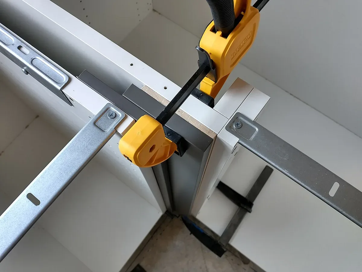 Cabinet face frames clamped together with a bar clamp during installation, with drawer slide rails and mounting brackets visible inside the boxes