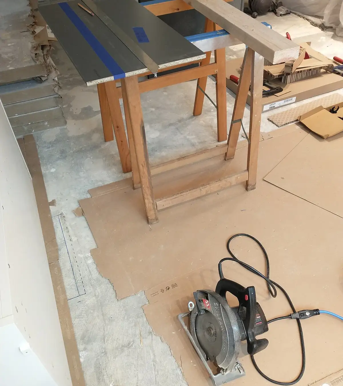 A circular saw, sawhorses and a cut panel on a kitchen job site with the floor papered and cabinets part installed
